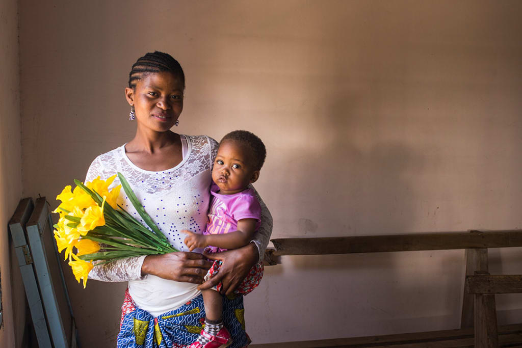 Neema, a Ugandan mother, stands inside a building holding her daughter, Tumaina and bouquet of yellow flowers.